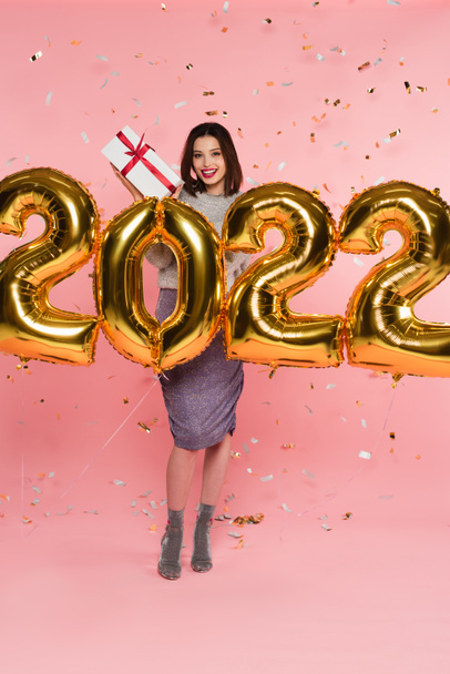 Smiling woman holding present near balloons in shape of 2022 and confetti on pink background - Fotografie, Obrázek