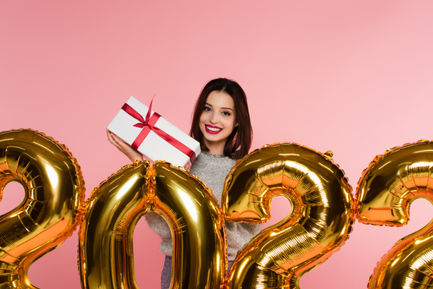 Happy woman holding present near balloons in shape of 2022 numbers isolated on pink  - Фото, изображение