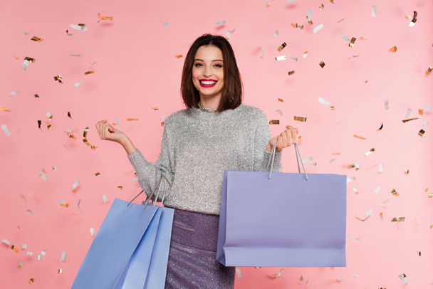Stylish woman in sweater holding shopping bags under confetti on pink background - Fotoğraf, Görsel