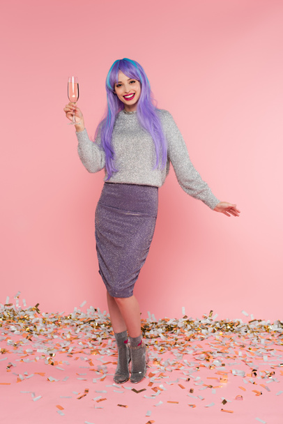 Happy stylish woman with dyed hair holding champagne near confetti on pink background - Fotoğraf, Görsel