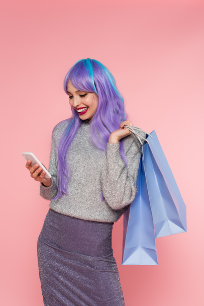 Glückliche Frau mit gefärbten Haaren nutzt Smartphone und hält Einkaufstüten isoliert auf rosa  - Foto, Bild