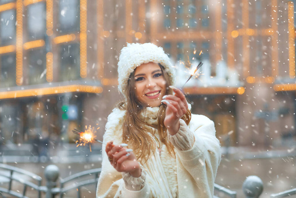 Pozitív fiatal nő visel fehér kötött kalap és kabát szórakozás csillagszórók a hóesés alatt - Fotó, kép