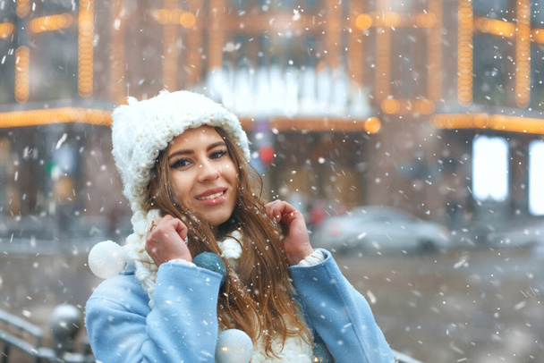 Mulher morena impressionante usa gorro de malha e casaco andando na cidade decorado com guirlandas durante a queda de neve. Espaço para texto - Foto, Imagem