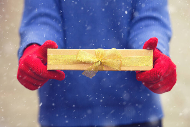 Woman wears a blue sweater and red winter gloves holding a gift box with a bow during the snowfall - Photo, Image