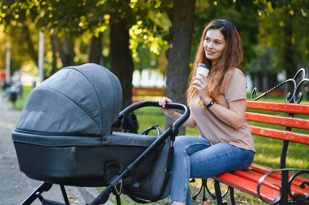 Stylish, young mother is sitting on a bench with a baby carriage in a park - Foto, Imagem