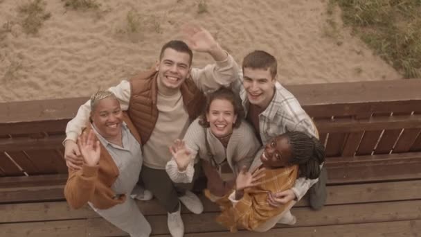 曇り空の日にビーチハウスのバルコニーに立つアフリカ人女性と白人女性と男性のトップビュー,抱擁,笑顔とカメラに手を振って - 映像、動画