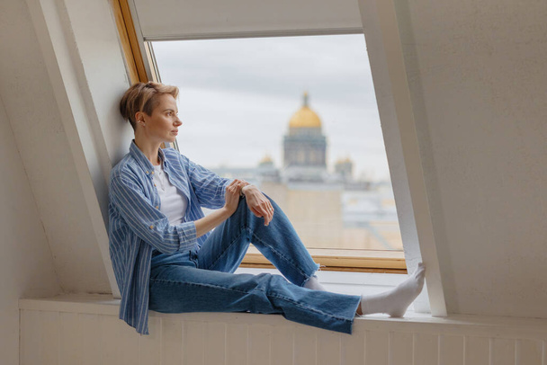a young and beautiful European woman with short blonde hair is sitting on the windowsill.  - Foto, Imagem