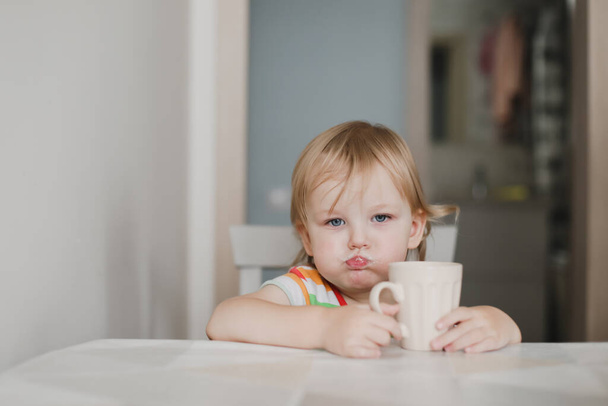 funny little girl drinking milk sitting in the kitchen - Zdjęcie, obraz
