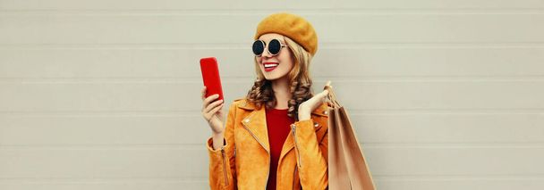 Autumn portrait of happy smiling young woman taking a selfie by smartphone with shopping bags wearing an yellow jacket, beret on gray background - Foto, Imagem