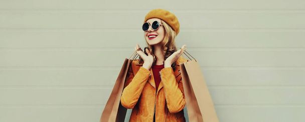 Jesienny portret pięknej uśmiechniętej młodej kobiety z torbami na zakupy w żółtej kurtce, beret na szarym tle - Zdjęcie, obraz