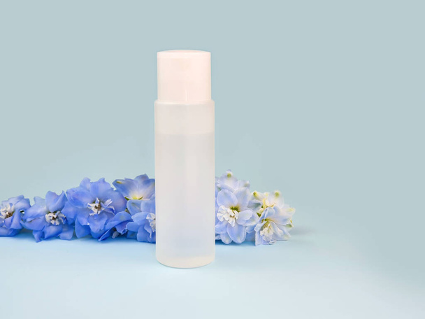 Fles met helder micellair water en blauwe delphiniumbloem op een blauwe achtergrond. Lotion voor het reinigen van de huid van cosmetica, make-up. Vloeibaar schoonheidsproduct in witte plastic verpakking, model, vooraanzicht - Foto, afbeelding