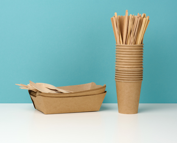 stack of brown disposable paper cups on a white table, wooden forks and knives, blue background. Utensils for takeaway drinks, zero waste - Foto, imagen