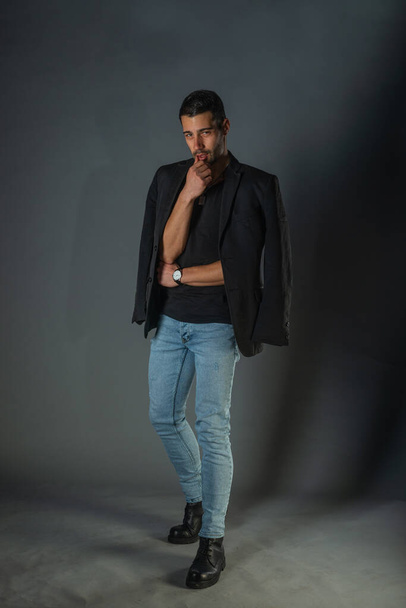 Voller Länge trendiger junger Mann in schwarzem Blazer und Jeans vor grauem Hintergrund - Foto, Bild
