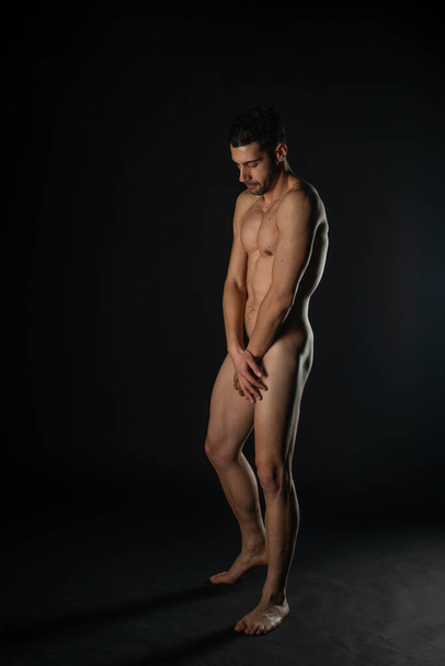 Full-length picture in a dark studio of a handsome naked man posing on the side - Фото, зображення