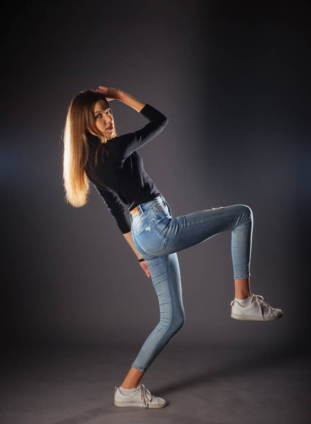 Side full-body portrait of a young female model in black top and jeans posing with one leg in front - 写真・画像