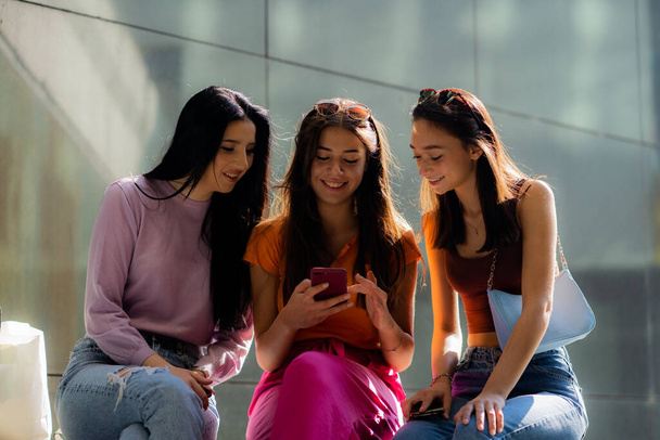Three attractive and beautiful girl friends are smiling at something on the phone while sitting - Photo, Image