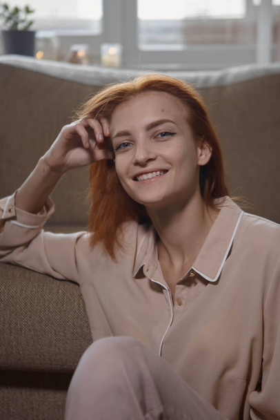 Caucasian young good looking woman with red hair sitting on the floor near the sofa in room and Woman looking at camera, smiling and laughing. Joy, warmth, happiness. - Photo, image