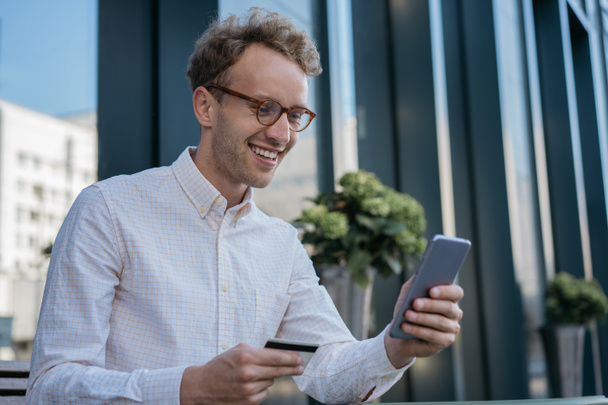 Young smiling businessman holding credit card, using mobile app for online shopping. Happy handsome freelancer check card balance, receive payment. Successful business concept - Foto, imagen