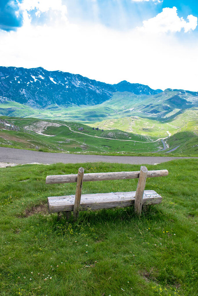 Dřevěná lavička na zeleném kopci na pozadí pohoří Durmitor National Park a silnice, která je nejvyšší silniční průsmyk v Černé Hoře. Krásná letní oblačná krajina. - Fotografie, Obrázek