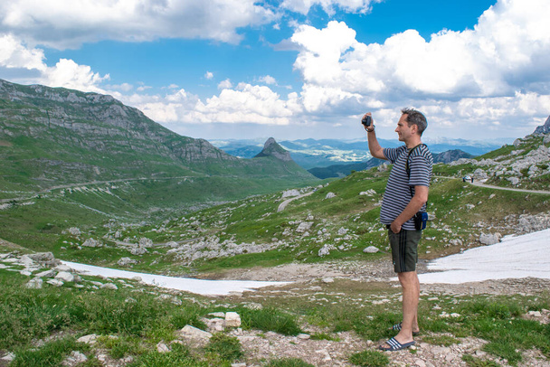 Man Tourist macht ein Foto von der schönen Sommerlandschaft. Berggipfel, grüne Hügel mit Schnee und wolkenloser blauer Himmel im Durmitor-Nationalpark. Höchster Straßenpass in Montenegro. - Foto, Bild