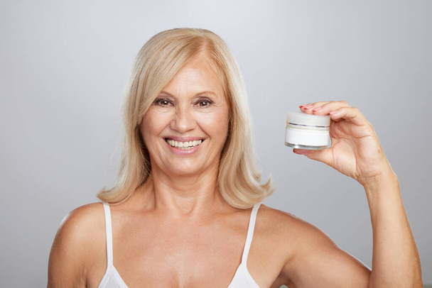 A happy senior blond woman holding anti-age creme in hand and looking at the camera. - Valokuva, kuva