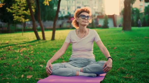 Joogaa puistossa. Keski-ikäinen nainen lootuksessa poseeraa joogamatolla vihreällä ruoholla. Rauhallisuuden ja meditaation käsite. Joogaa ulkona. Vanhempi nainen meditoi ja käyttää jooga lootus asema luonnossa - Materiaali, video