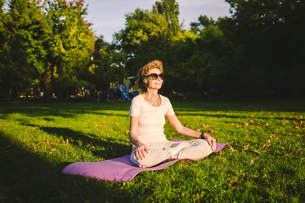 Senior vrouw mediteren en oefenen yoga lotus positie buiten. Ederly vrouw doen stretching oefeningen op yoga mat in park op groen gazon bij zonsondergang. Ik voel energie. Concept rust en meditatie. - Foto, afbeelding