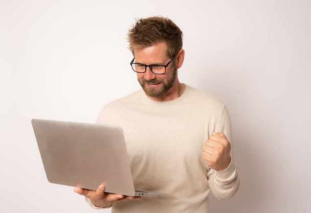 Studio portrait of a young happy man in a casual sweater with a laptop making winner gesture on a white background - 写真・画像