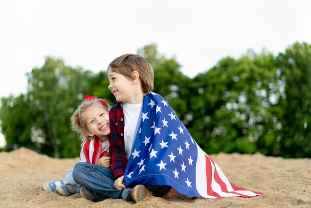 brother and sister on the shore, wrapped in the American flag, happy children laughing, girl and boy concept of patriotism and the celebration of U.S. independence. U.S. Veterans Day. - Photo, Image