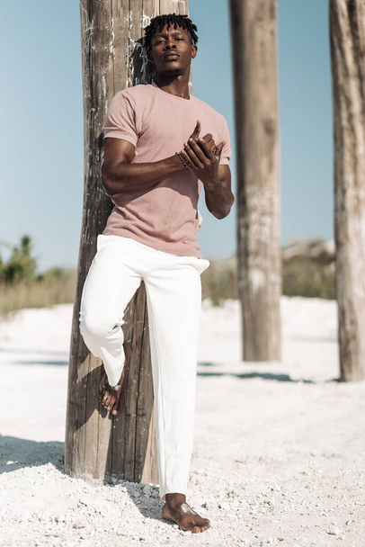 African American man with African hair in a pink T-shirt and white pants, looking to the side near wooden pillars, a man in an exotic desert, sand dunes around, Fashion model concept, mens clothing - Photo, image
