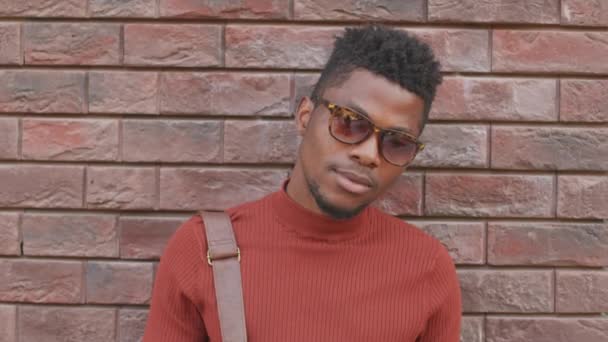 Mittleres Nahaufnahme-Porträt mit Slowmo eines hübschen jungen afroamerikanischen Mannes mit rotem Rollkragen und trendiger Sonnenbrille, der im Freien vor einer Backsteinmauer für die Kamera posiert - Filmmaterial, Video