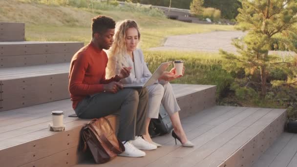 Slowmo facada tiro de jovem casal de negócios inter-raciais em casualwear inteligente olhando para laptop e ter uma conversa sentada com tomar café nas escadas no parque no dia ensolarado - Filmagem, Vídeo