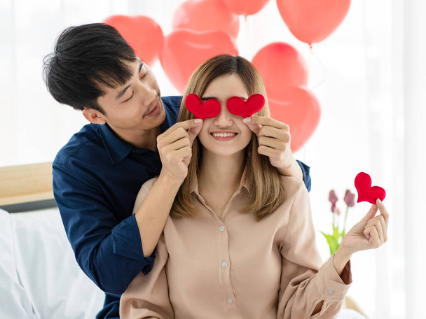 Asiatischer Mann hält Herzen in der Nähe von Augen freudiger Freundin, während er sich am Valentinstag zu Hause auf dem Bett ausruht - Foto, Bild