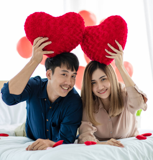Positive Asian man and woman looking at camera with smile and demonstrating heart shaped pillows while resting on bed during Saint Valentine Day celebration. Love and relationship of teens lovers concept. - Photo, Image