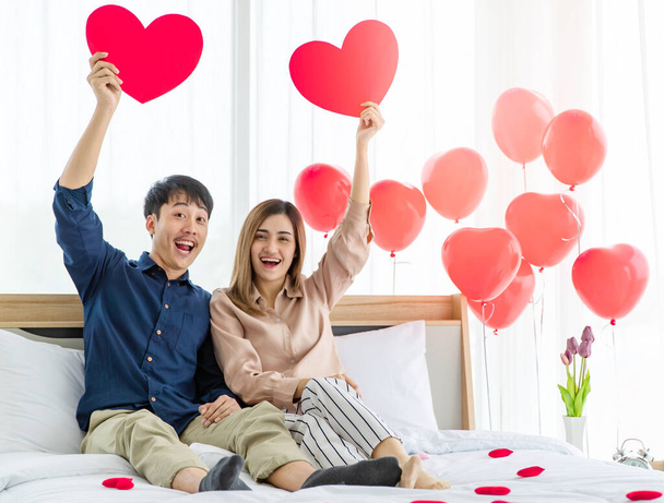 optymistyczny Azji mężczyzna i kobieta para miłośników patrząc w aparat z uśmiechem i podnosząc ręce z papieru serca podczas odpoczynku na łóżku w święte Walentynki - Zdjęcie, obraz