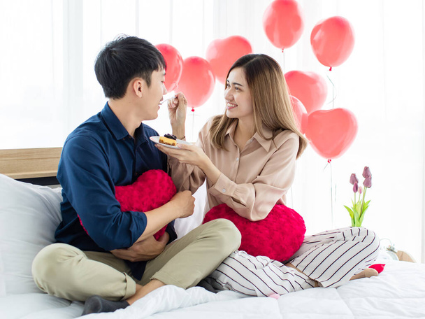 Szczęśliwy para miłośników, Azjatki dzielenie słodkie ciasto z mężczyzną za pomocą laptopa podczas odpoczynku na łóżku na Saint Valentine Day - Zdjęcie, obraz