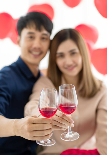 Wesoły azjatycki mężczyzna i kobieta uśmiechają się, pijąc kieliszki wina i proponując toast podczas obchodów Walentynek w domu. Selektywne skupienie się na okularach. - Zdjęcie, obraz