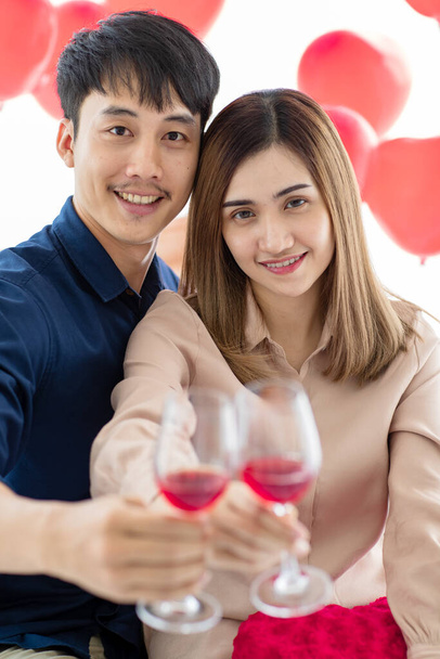 Alegre asiático hombre y mujer sonrisa mientras tintineo vasos de vino y proponer tostadas durante la celebración del día de San Valentín en casa. Enfoque selectivo en las caras de la pareja. - Foto, Imagen