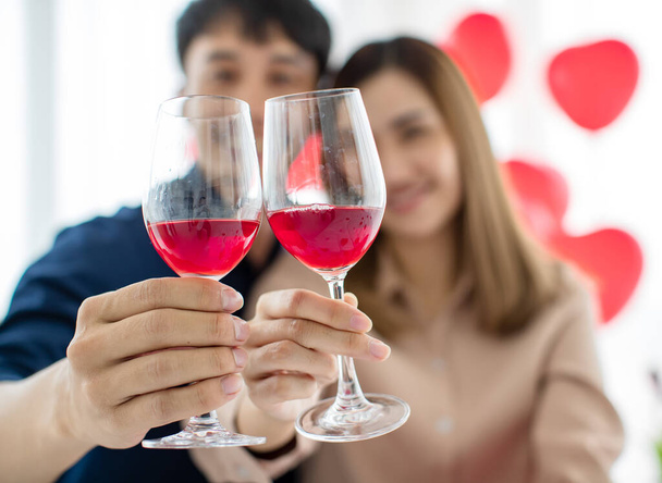 Fröhliche asiatische Männer und Frauen lächeln, während sie Gläser mit Wein klappern und während der Valentinstagsfeier zu Hause anstoßen. Selektiver Fokus auf Brillen. - Foto, Bild