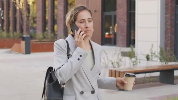 Средневековый словесный снимок серьезной кавказской бизнесвумен в элегантном брючном костюме, прогуливающейся по центру города с отнятой чашкой кофе в руках и разговаривающей по телефону с деловым партнером - Кадры, видео