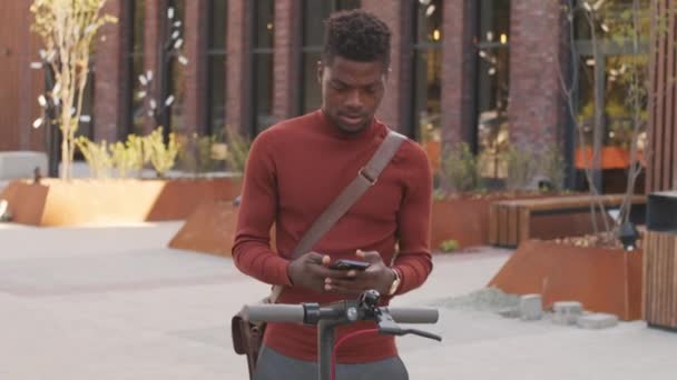 若いですハンサムなアフリカ系アメリカ人の男のティルトアップ中低速ショットスマートカジュアルウェアで彼のスマートフォン上のアプリを使用して電動キックスクーターを設定ビジネス地区で屋外に立って - 映像、動画