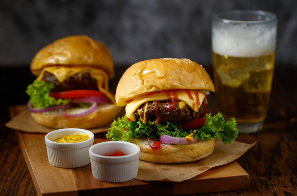 Крупный план готового к употреблению домашнего говяжьего гамбургера со свежими овощами с двумя цветными догонялками и стаканом холодного пива со льдом на заднем плане. - Фото, изображение