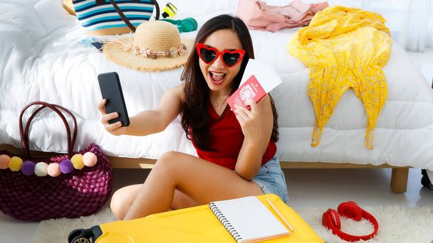 Jovem feliz e excitar asiático viajante feminino se preparando para uma viagem de férias de verão com sua mala amarela brilhante usando óculos de sol em forma de coração tirando foto selfie com seu passaporte. - Foto, Imagem