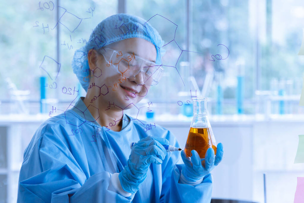 Portret foto van Aziatische vrouwelijke wetenschapper in beschermende pak veiligheidsbril en rubber handschoenen staan lachend gebruik marker pen labeling op Erlenmeyer fles overlappen met chemische formule op de voorgrond. - Foto, afbeelding