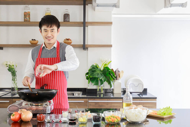 Retrato de joven marido guapo asiático lleva delantal de raya roja de pie sonriente mira a la cámara vertiendo ajo picado de vidrio en la cena de cocina en la estufa eléctrica en la encimera de la cocina. - Foto, Imagen