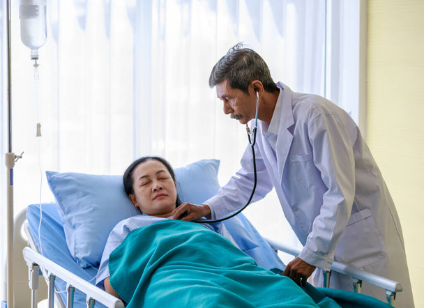 Starý senior starší asijské šedé vlasy a knír lékař v bílém laboratorním kabátu pomocí stetoskopu monitor poslouchat srdeční tep pacientky, která spí na nemocničním lůžku s normálním solným roztokem kapání. - Fotografie, Obrázek