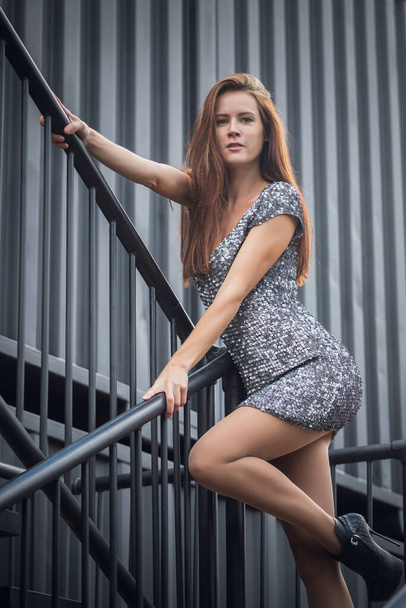 Retrato de cerca al aire libre de una joven hermosa con un vestido brillante posando en las escaleras contra el telón de fondo de un contenedor industrial Concepto de caminata de otoño. - Foto, imagen