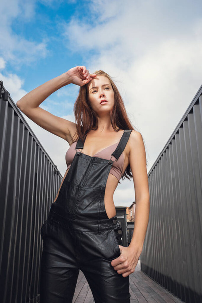 Creative portrait of a red-haired woman in  leather overalls and top . Design concept. Romantic portrait of young woman posing in urban area - Fotografie, Obrázek