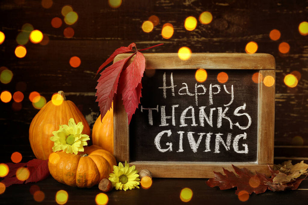 Chalkboard with text HAPPY THANKSGIVING and autumn decor on dark wooden background with blurred lights - Photo, Image