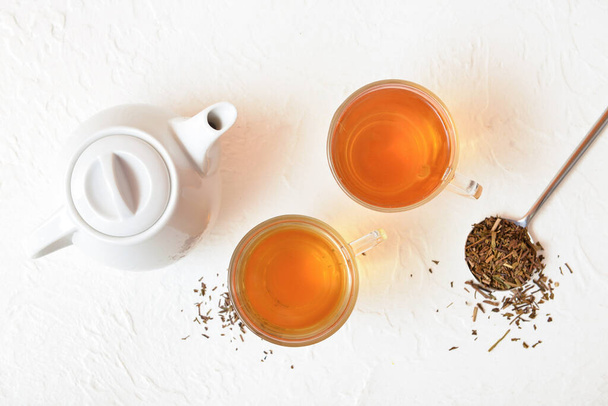 Skleněné šálky chutného hojicha čaje, hrnce a suché listy na bílém pozadí - Fotografie, Obrázek